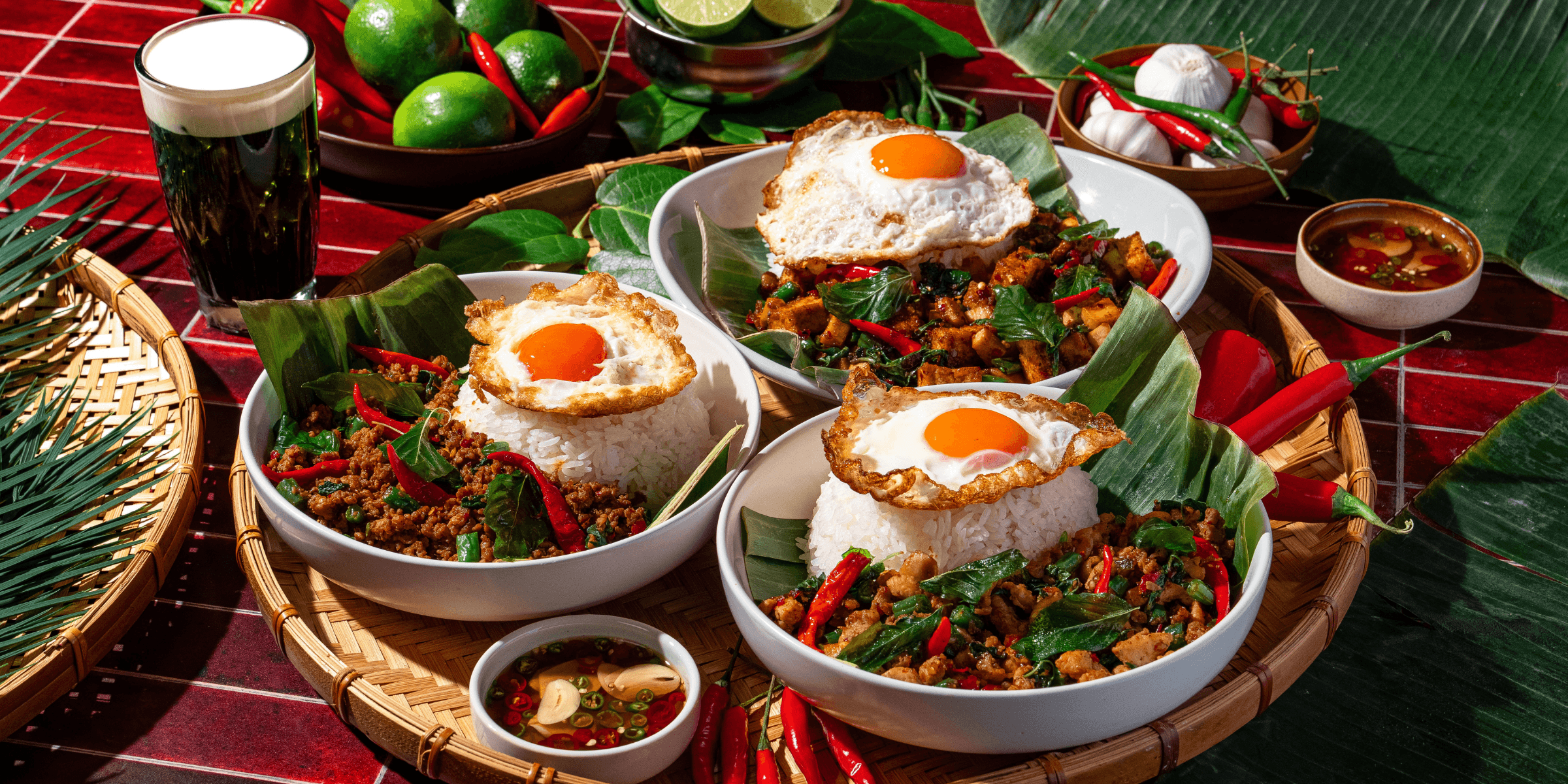 Holy Heat Pad Kra Pow bowls with spicy Thai basil stir-fry, white rice, and crispy fried eggs, surrounded by fresh chilies, limes, garlic, and Thai-inspired ingredients.
